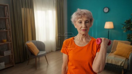 Foto de Retrato de una anciana de pelo gris haciendo ejercicios de fitness con pesas en casa de cerca. Una mujer madura bombea sus bíceps, entrena sus tríceps, mantiene sus músculos del brazo en buena forma. Deportes para - Imagen libre de derechos