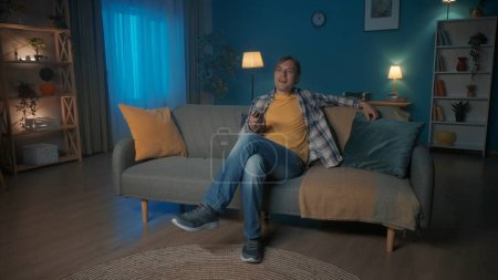 Téléchargez les photos : Un homme s'assoit sur le canapé le soir et regarde la télévision. Un homme change de canal avec une télécommande - en image libre de droit