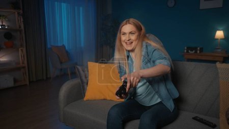 Foto de Una mujer juega un juego consular con un controlador de juego mientras está sentada en el sofá en casa por la noche. Mujer concentrada presiona joystick, nervioso - Imagen libre de derechos