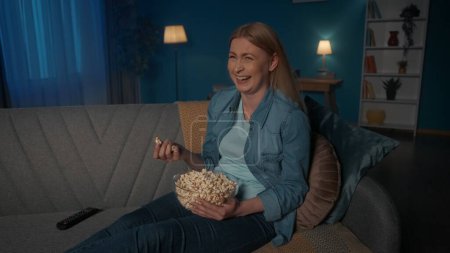 Foto de Retrato de una joven riendo con un plato de palomitas de maíz en sus manos de cerca. Una mujer se sienta en un sofá y ve la televisión. Película nocturna viendo, descansar en casa - Imagen libre de derechos