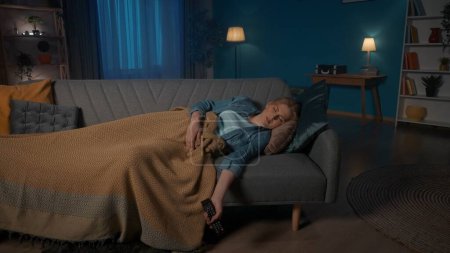 Foto de Una mujer cansada y somnolienta se quedó dormida mientras veía la televisión. Una mujer duerme, cubierta con una manta, acostada en el sofá de la sala de estar, sin soltar el mando a distancia - Imagen libre de derechos