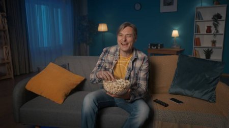 Foto de Un hombre se sienta en el sofá por la noche, come palomitas de maíz y ve la televisión. Un hombre ve una comedia, un programa de comedia, se ríe alegremente - Imagen libre de derechos