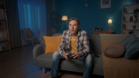 Photo for A man plays a consular game with a game controller while sitting on the couch at home in the evening. A man often presses the joystick, gets nervous - Royalty Free Image