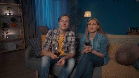 Téléchargez les photos : Un jeune couple est assis sur un canapé avec des verres de vin rouge et regarder la télévision. Un homme et une femme regardent un film avec intérêt, une vidéo touchante. Week-end concept de loisirs - en image libre de droit