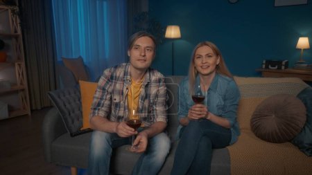 Foto de Una joven pareja está sentada en un sofá con copas de vino tinto y viendo la televisión. Un hombre y una mujer están viendo una película con interés, un video conmovedor. Concepto de ocio fin de semana - Imagen libre de derechos