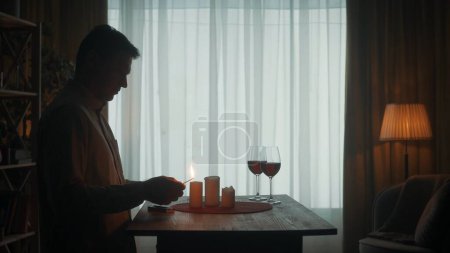 Photo for The man is getting ready for a date. The dark silhouette of a man lights candles with matches. Candles and glasses with red wine on the table - Royalty Free Image