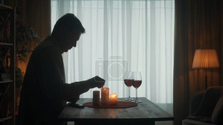 Foto de El hombre se está preparando para una cita. La silueta oscura de un hombre enciende velas con fósforos. Velas y copas con vino tinto sobre la mesa - Imagen libre de derechos