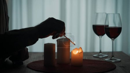 Photo for Romantic evening by candlelight. Hands of a man lighting candles with matches close up. Burning candles and glasses with red wine on the table - Royalty Free Image