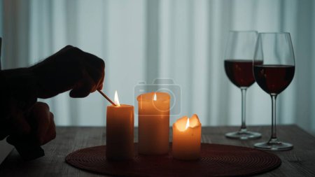 Photo for Romantic evening by candlelight. Hands of a man lighting candles with matches close up. Burning candles and glasses with red wine on the table - Royalty Free Image
