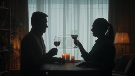 Téléchargez les photos : Le couple passe la soirée avec du vin rouge aux chandelles, on dit des toasts, on lève les verres, on parle. Silhouettes sombres d'un homme et d'une femme dans le salon à la table. Soirée romantique - en image libre de droit