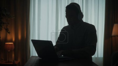 Téléchargez les photos : Silhouette sombre d'un homme dans un casque sans fil à une table devant un ordinateur portable de près. Un homme travaille sur un ordinateur portable, écoute de la musique - en image libre de droit