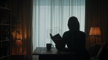 Téléchargez les photos : Une femme lit un livre assis à une table avec une tasse de café, du thé dans la pièce sur le fond de la fenêtre. Silhouette sombre d'une femme avec un livre ouvert à la main - en image libre de droit