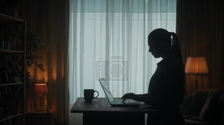 Téléchargez les photos : Vue latérale d'une silhouette sombre d'une femme à une table devant un ordinateur portable. Une femme travaille sur un ordinateur portable avec une tasse de café, jouer à un jeu en ligne - en image libre de droit