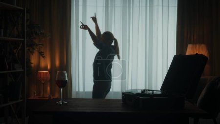 Foto de Grabador vintage con un disco de vinilo y una copa de vino tinto en la mesa de cerca. Mujer despreocupada baila alegremente sola cerca de la ventana. Silueta mujer bailarina en la sala de estar - Imagen libre de derechos