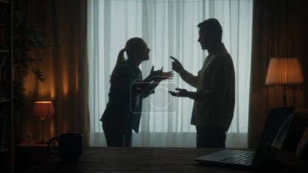 Foto de Un hombre y una mujer discuten en la sala de estar cerca de la ventana. Una silueta de una pareja discutiendo sobre el hecho de que el hombre pasa demasiado tiempo en la computadora portátil - Imagen libre de derechos