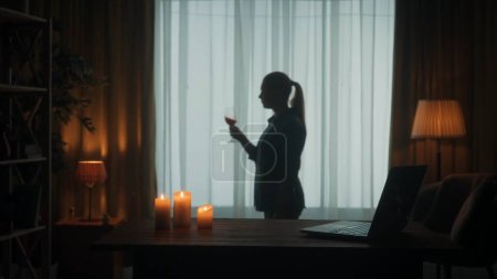 Photo for A young woman enjoys wine after a hard day at work. Burning candles and a laptop are on the table, a woman shakes a glass of wine. Blurred dark silhouette of a woman near the window - Royalty Free Image
