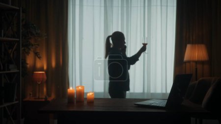 Foto de Una mujer habla por teléfono y sostiene una copa de vino. Encender velas y un portátil están sobre la mesa, una mujer examina una copa de vino. Silueta oscura borrosa de una mujer cerca de la ventana - Imagen libre de derechos