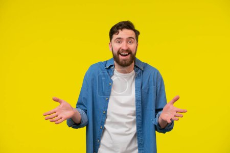 Photo for A portrait of a bearded man with spreaded open palms sideways. Looks with joy and surprise. Smiling happily with eyes and mouth wide open. Standing against yellow background, looking at the camera. - Royalty Free Image