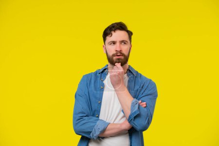 Photo for A portrait of a young adult bearded man looking away and thinking, touching his chin, crossing arms. Full of thought and concentrated. Isolated on the yellow background. - Royalty Free Image