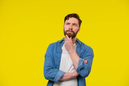 A portrait of a young adult man wearing casual clothes, looking up and thinking. Dreaming about something, touching his chin. Isolated on the yellow background.