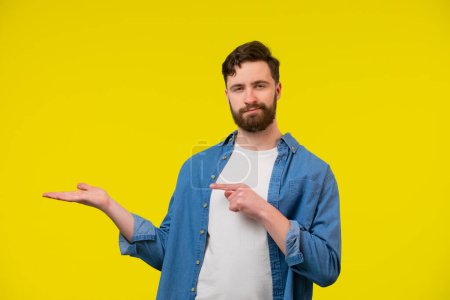 Photo for Portrait of a young man in blue shirt holding his arm up with opened palm, pointing his finger and looking at the camera. Space for objects. Isolated on yellow background. - Royalty Free Image