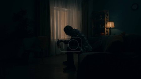 Foto de Un joven devastado se sienta en una habitación en la oscuridad con una botella de alcohol. Alcoholismo. Desesperanza. Tratamiento de la adicción al alcohol. El concepto de salud mental - Imagen libre de derechos