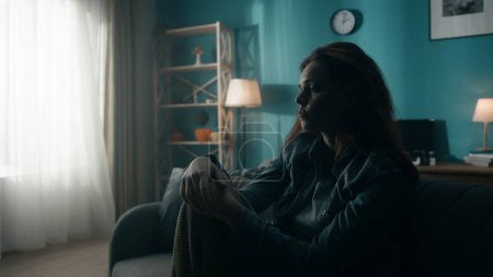 Foto de Una joven devastada y solitaria se sienta en un sofá en una habitación oscura de cerca. Una mujer piensa desesperadamente en problemas personales, experimentando una crisis. Desesperanza. Concepto de salud mental - Imagen libre de derechos