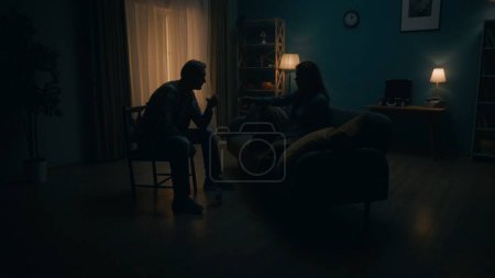 Foto de Una pareja se sienta con vasos de alcohol en una habitación en la oscuridad. Un hombre y una mujer se sientan uno frente al otro, entre ellos una botella de alcohol. La pareja escapa de los problemas con la ayuda de - Imagen libre de derechos
