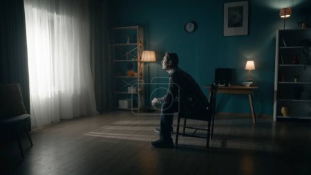 Photo for A man sits on a chair in the middle of the room and looks at the light in the window. Side view of a lonely man sitting in a dark room. Hope concept, mental health - Royalty Free Image