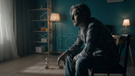 Photo for A man sits in a dark room on a chair close up, in front of a window from which light streams. The man looks into the light, holds a glass of alcohol in his hand, there is an almost empty bottle on the - Royalty Free Image