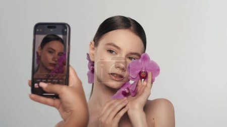Téléchargez les photos : Gros plan d'un beau modèle au maquillage naturel posant pour une séance photo sur un smartphone, tenant une fleur d'orchidée, souriant heureux. Concept de publicité sur les médias sociaux. - en image libre de droit