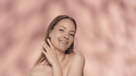 Téléchargez les photos : Joyeux sourire jeune femme dans le studio sur un fond rose. Une femme touche sa main avec une douce manucure au cou, appréciant la douceur de la peau. Concept beauté. Beauté naturelle - en image libre de droit