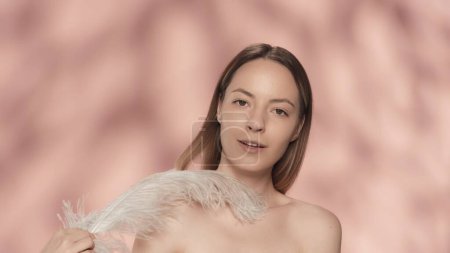 Photo for A woman touches her bare shoulders with a white feather. A young woman demonstrates smooth and soft skin in the studio on a pink background. The concept of beauty, cosmetology, care - Royalty Free Image