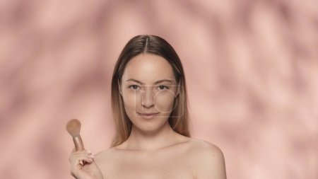 Foto de Retrato de una mujer seminude con un cepillo de maquillaje en el estudio sobre un fondo rosa de cerca. Cosmética facial. Piel fresca y maquillaje natural. El concepto de belleza, cosmetología, cuidado de la piel - Imagen libre de derechos