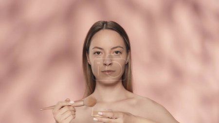 Téléchargez les photos : Femme avec un pinceau de maquillage et un pot de poudre en studio sur un fond rose. Contouring, application de poudre, rougeur. Le concept de beauté, cosmétologie, soin de la peau - en image libre de droit