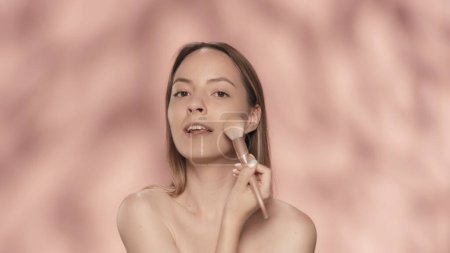 Téléchargez les photos : Seminude femme utilise un pinceau de maquillage en studio sur un fond rose de près. Une femme fait du contouring, applique de la poudre, rougit. Cosmétiques pour le visage. Peau fraîche et maquillage naturel. Le concept de beauté - en image libre de droit