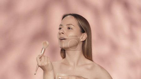Téléchargez les photos : Seminude femme utilise un pinceau de maquillage en studio sur un fond rose de près. Une femme fait du contouring, applique de la poudre, rougit. Cosmétiques pour le visage. Peau fraîche et maquillage naturel. Le concept de beauté - en image libre de droit
