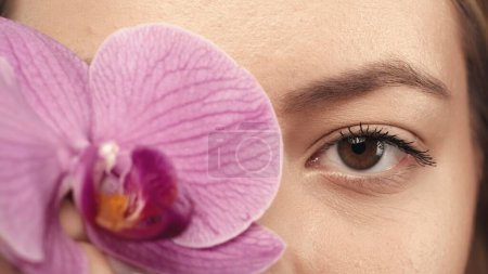 Foto de Hermosos ojos de una joven de cerca. Una mujer cubre su ojo con una flor de orquídea. Buena visión y cuidado ocular. Lentes de contacto. Belleza femenina natural. Línea cosmética para el cuidado del contorno de ojos - Imagen libre de derechos