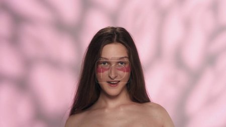Téléchargez les photos : Portrait d'un beau modèle de brune caucasienne. Gros plan d'une fille avec des patchs roses sous les yeux, souriant à la caméra. Concept publicitaire de soins de la peau pour les yeux. - en image libre de droit