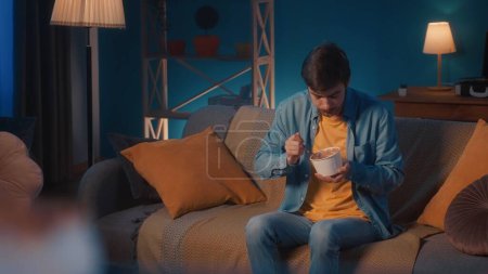 Photo for A man greedily eats ice cream from a bucket while sitting on the sofa in the living room - Royalty Free Image