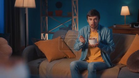 Foto de Un hombre se sienta en un sofá en la sala de estar y come helado con una cuchara de un cubo. Un hombre con una cucharada llena de helado mira hacia adelante con sorpresa - Imagen libre de derechos