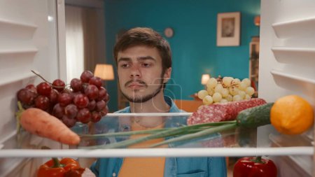 Photo for A young frustrated man looks inside the refrigerator. A man examines healthy products on the shelves with a disappointed look, there is nothing to choose to eat - Royalty Free Image