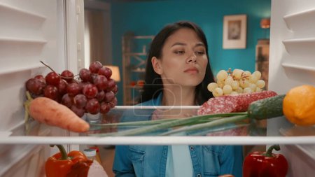 Photo for Puzzled Asian woman looks inside the refrigerator. A woman examines healthy products on the shelves with a disappointed look, there is nothing to choose to eat - Royalty Free Image