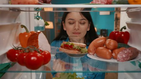 Photo for An Asian woman looks into the refrigerator, examines its contents. A woman, pulling up a plate with a sandwich, inhales its aroma with pleasure, closing her eyes. Choice between healthy and unhealthy - Royalty Free Image