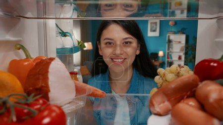 Foto de Retrato de una mujer asiática sonriente en el refrigerador abierto. Una mujer está satisfecha con la abundancia de alimentos en el refrigerador. Vista desde el interior del refrigerador - Imagen libre de derechos
