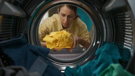 Photo for Adult woman in casual clothing with laundry basket opens the door of the washer and takes out the clean clothes. View from inside the washing machine. - Royalty Free Image