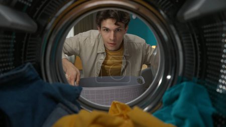Photo for Adult man in casual clothing with laundry basket opens the door of the washer and takes out the fresh clothes. View from inside the washing machine. - Royalty Free Image