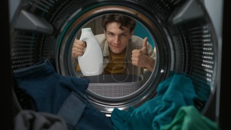 Photo for Adult man in casual clothing with laundry basket holding a bottle of softener and shows a thumbs up. View from inside the washing machine. - Royalty Free Image