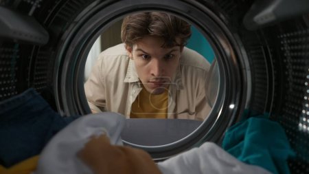 Photo for A portrait of a young adult man in casual clothing looking inside the washer drum full of clothes. View from inside the washing machine. - Royalty Free Image