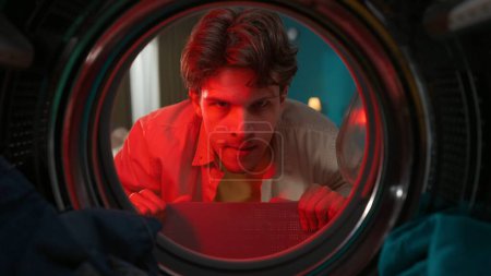 Photo for A portrait of a young adult man in casual clothing looking angry inside the washer drum full of clothes. Red light alert. View from inside the washing machine. - Royalty Free Image